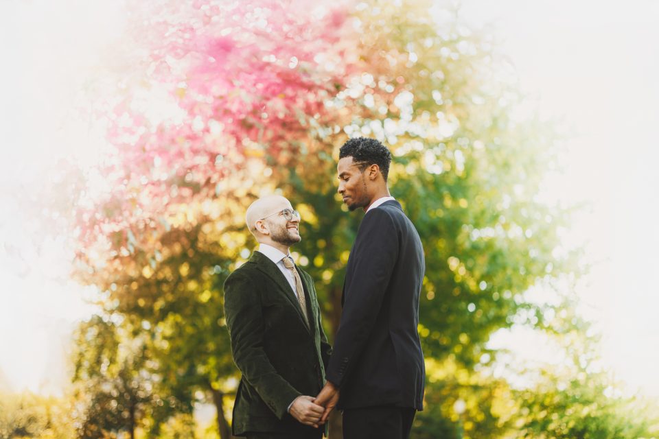 Two people stand holding hands outdoors in front of colorful autumn foliage, wearing formal jackets.