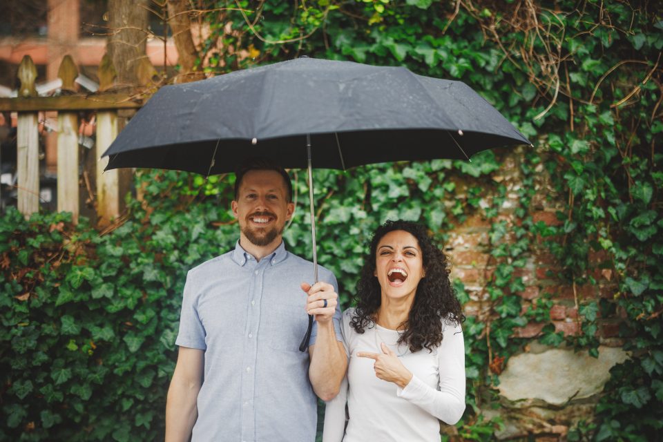 A man and woman stand under an umbrella in front of a wall with ivy. The woman is laughing and pointing at the man.