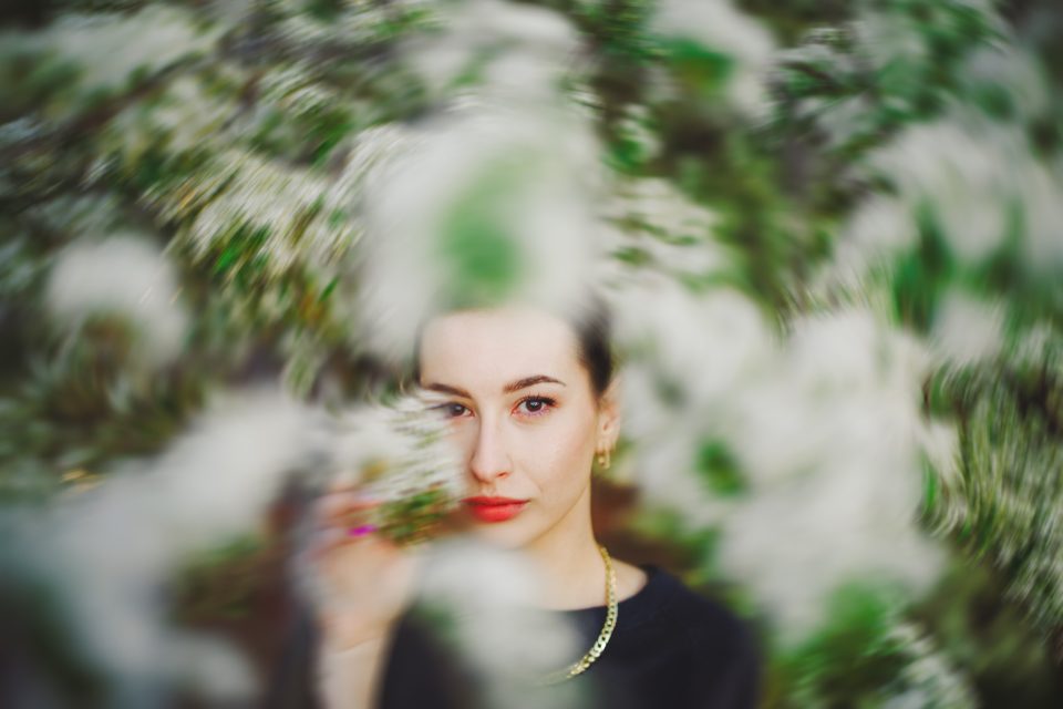 Woman with red lipstick and gold necklace peers through blurred white and green foliage, creating an artistic effect.
