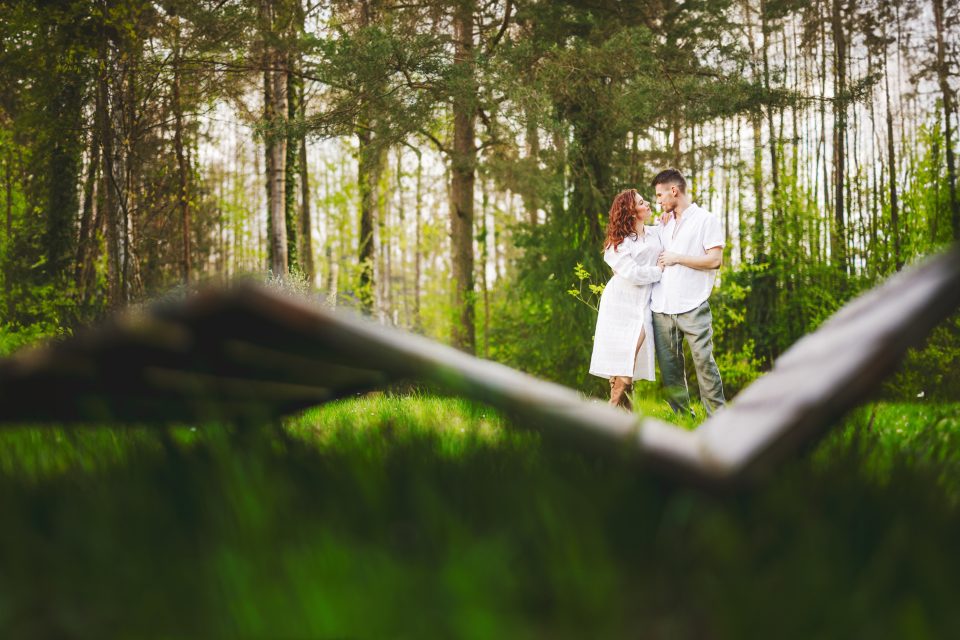 A couple embraces in a grassy forest clearing, surrounded by tall trees, with part of a wooden structure in the foreground.