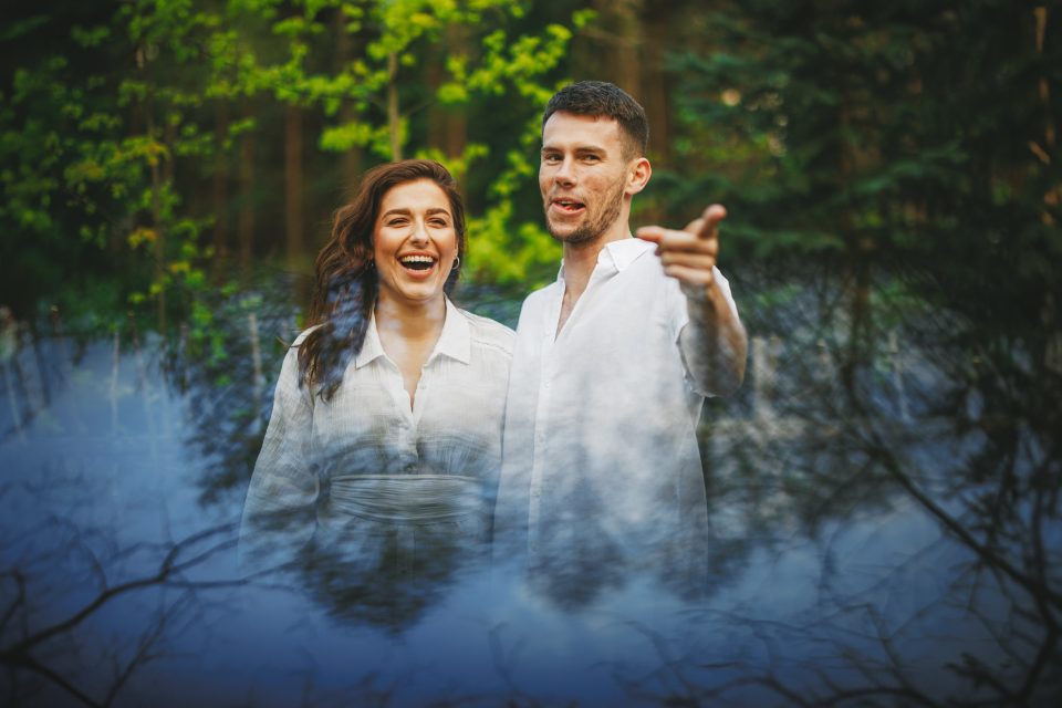 A smiling couple in white shirts stands outdoors, with the man pointing forward. The background is a blend of green foliage and a misty overlay.