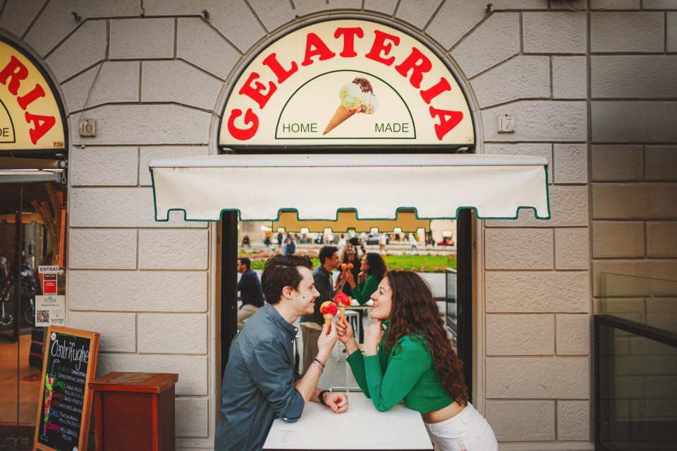 A couple shares ice cream at an outdoor table in front of a gelateria, under a sign reading "Gelateria Home Made." They smile, enjoying their treat.