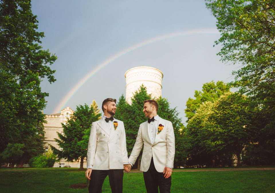Two people in white suits hold hands in a park with greenery and a rainbow in the background.
