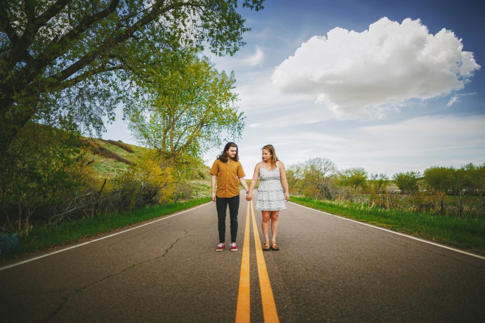 Two people holding hands, walking on a rural road lined with trees, under a cloudy sky.