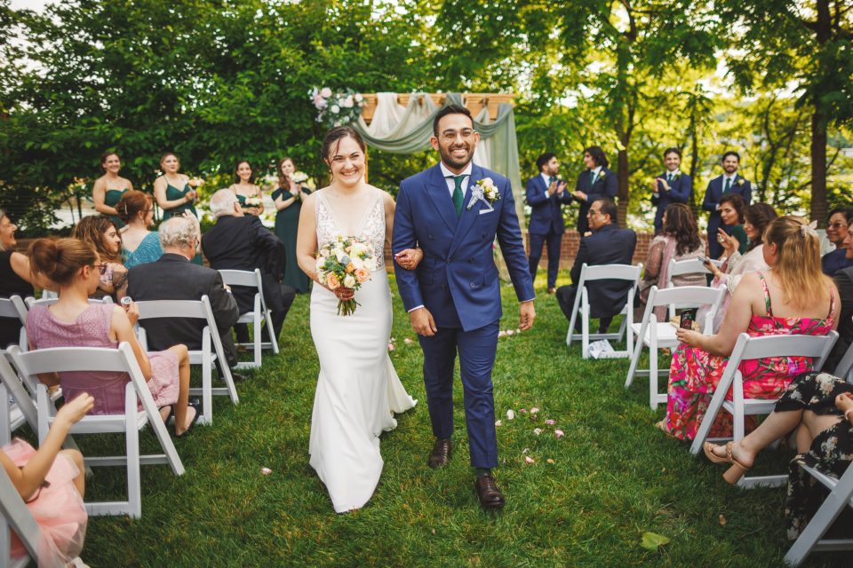 A bride and groom smile as they walk down the aisle outdoors, surrounded by seated guests and a wedding party.