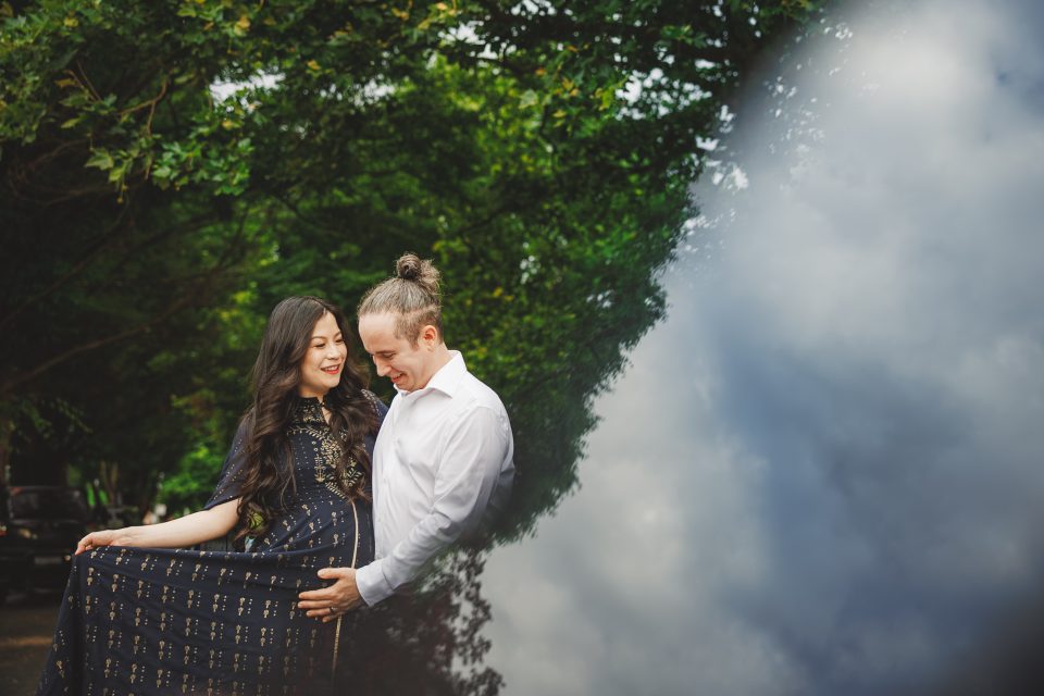 A couple stands closely outdoors, smiling. The woman holds her dress over her pregnant belly. Lush green trees are in the background, and there is a cloud reflection on the right side.