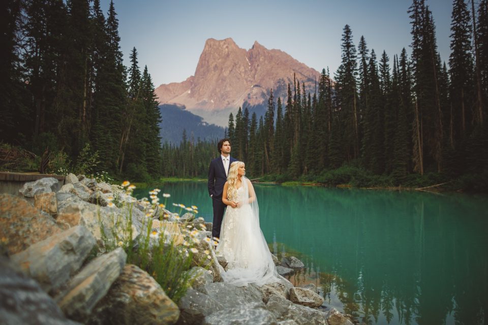 Bride and groom stand on rocky shoreline by a turquoise lake, surrounded by pine trees, with a mountain peak in the background.