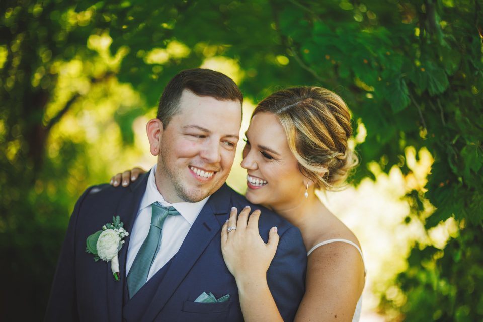 A couple in wedding attire smiles at each other under leafy green trees. The groom wears a blue suit; the bride is in a white dress.