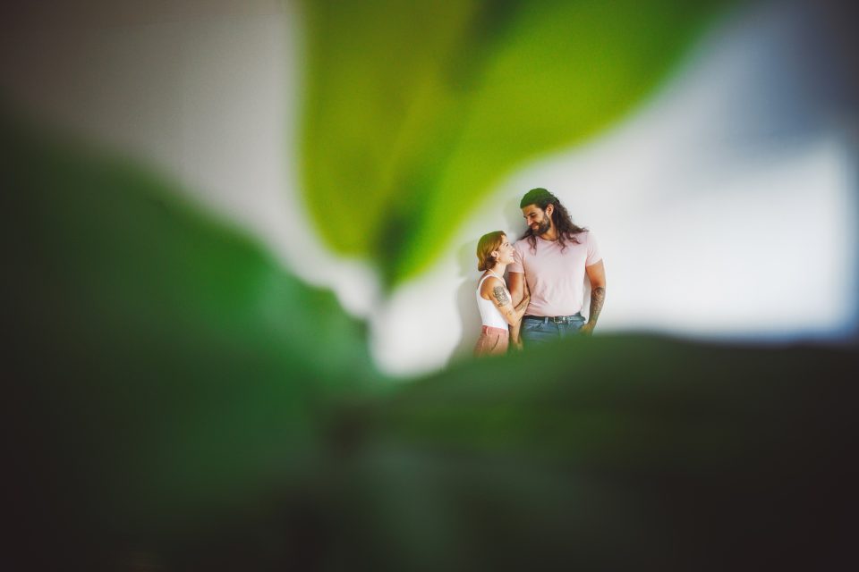 A couple stands close, smiling at each other, partially obscured by large green leaves in the foreground.