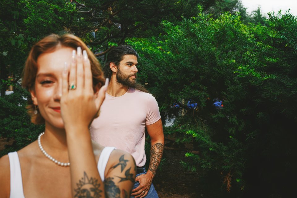 A person with tattoos covers their face with one hand, showing a ring, while another person stands in the background among greenery.