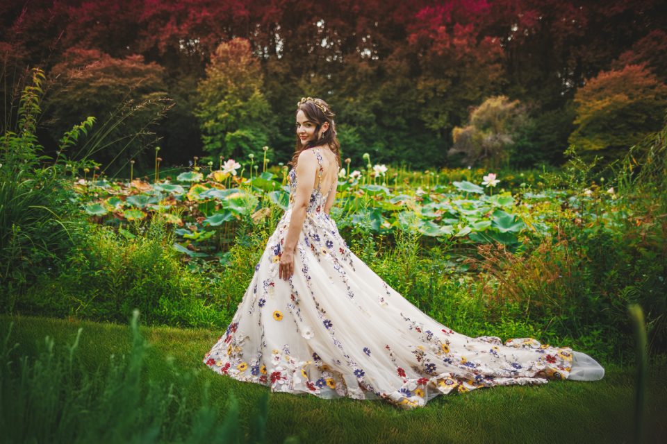 A woman in a floral gown stands on grass, surrounded by lush greenery and blooming flowers.
