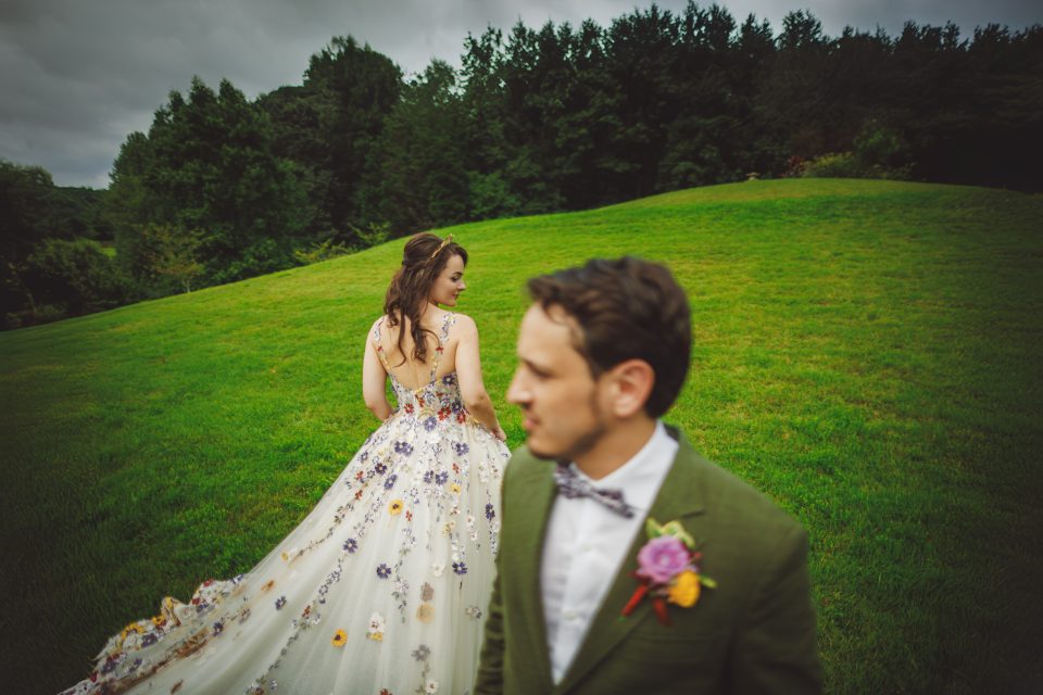 Bride in a floral gown stands on a green hill, looking over her shoulder. A man in a green suit with a flower boutonniere is in the foreground, slightly out of focus.