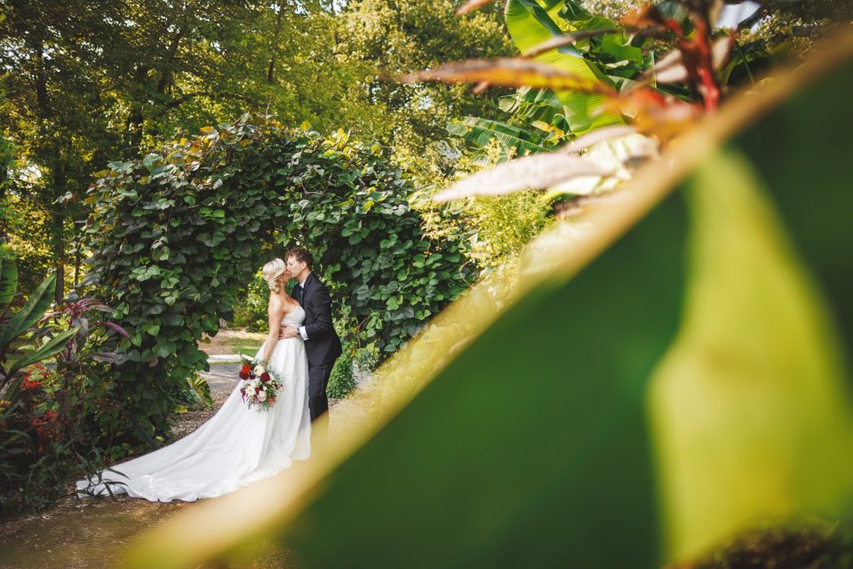 Bride and groom embrace in a lush garden with leafy arches and sunlight filtering through the trees.