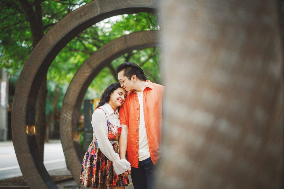 A couple stands closely, smiling, in front of large metal circular sculptures with trees in the background.