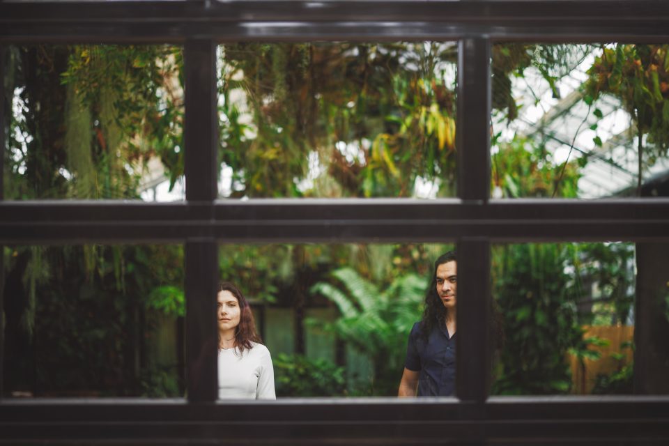Two people stand behind a framed glass structure in a lush greenhouse, surrounded by various plants and greenery.