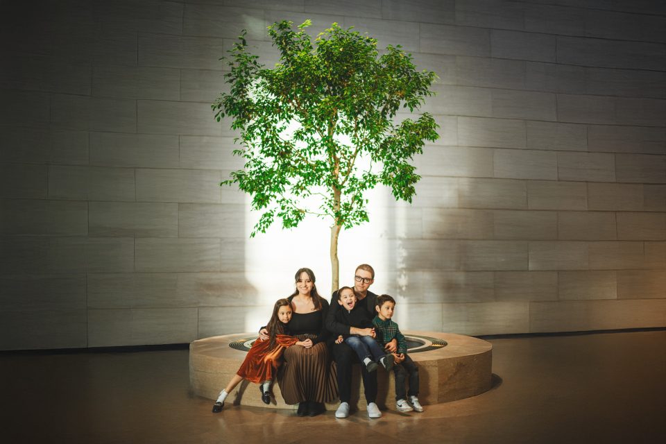 A family of five, with three children, sits on a circular stone bench in front of a large indoor tree, against a modern stone wall backdrop.