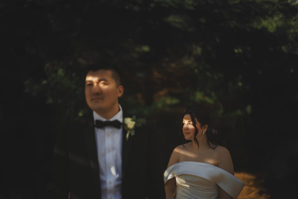 A couple stands outdoors, dressed formally; the man wears a tuxedo and the woman wears an off-shoulder dress. Dappled sunlight falls on them.