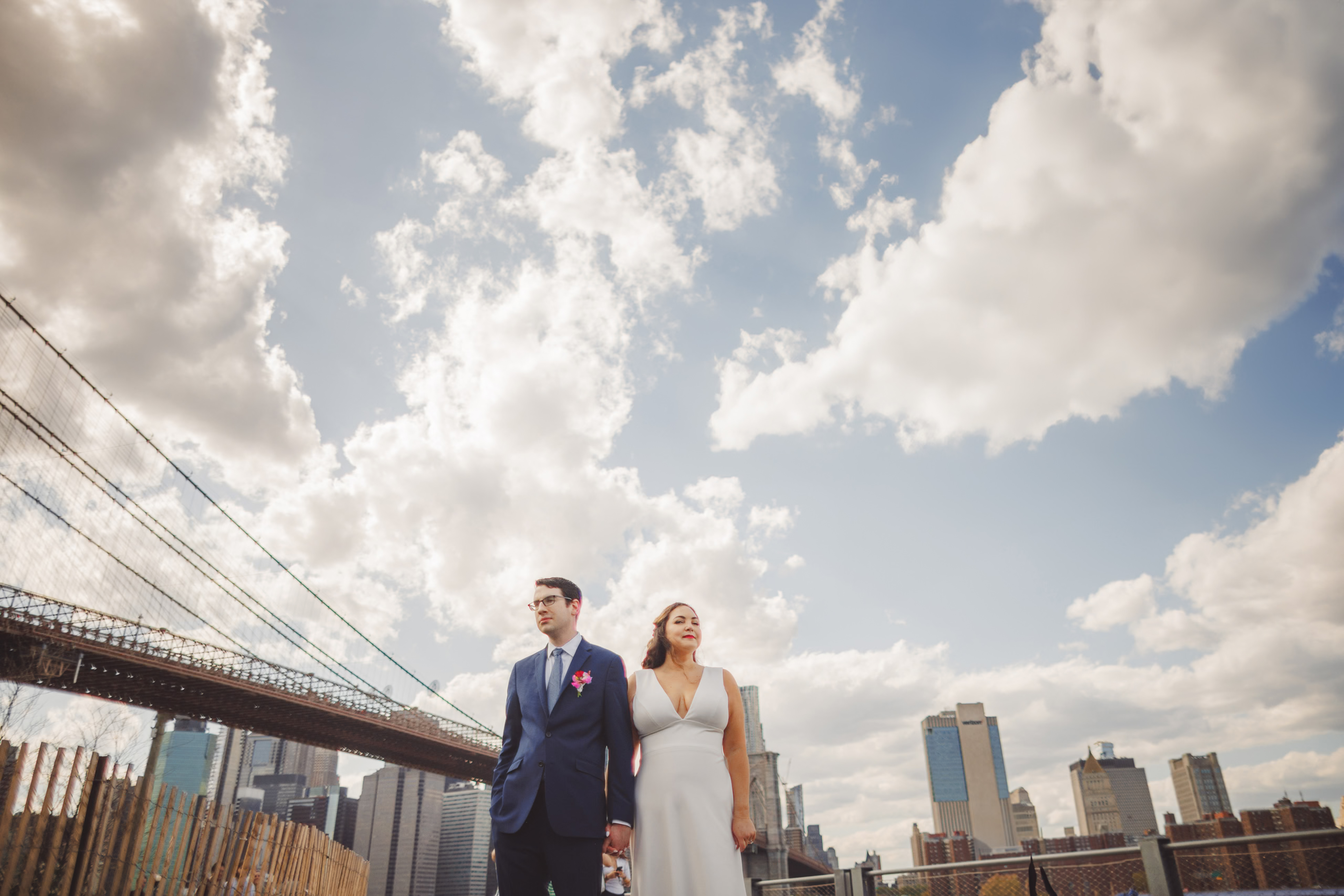 A couple in wedding attire stands near a railing, with the Brooklyn Bridge and New York City skyline in the background under a partly cloudy sky.
