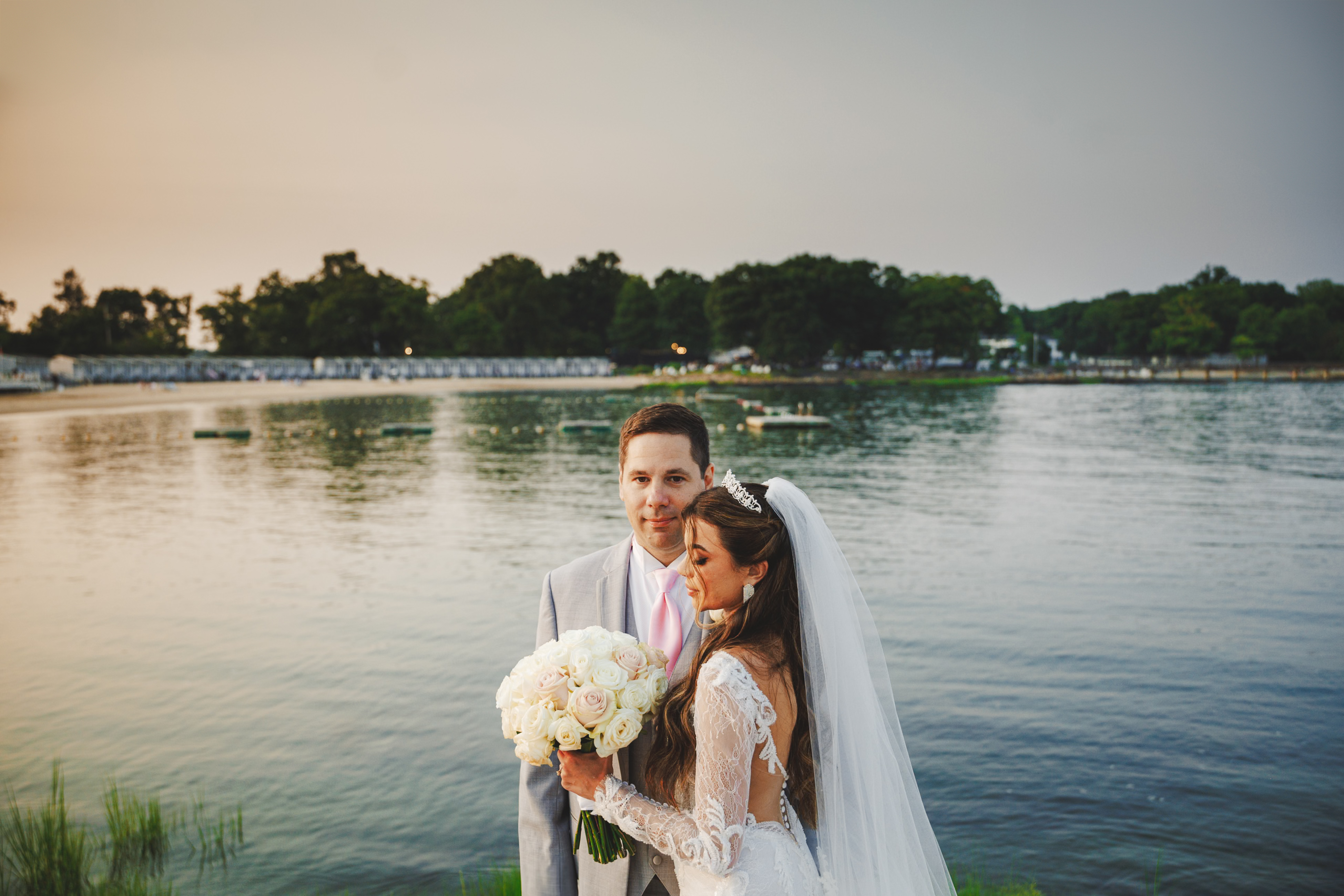 Bride and groom stand by a lake, with the bride holding a bouquet of white flowers. Trees and a cloudy sky are in the background.