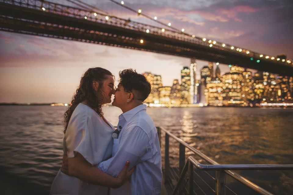 Two people embrace and look at each other on a waterfront at dusk, with a lit-up bridge and city skyline in the background.