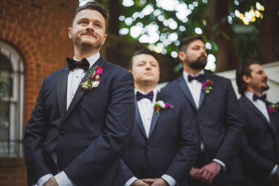 Four men in matching dark suits, white shirts, and bow ties stand outdoors, each wearing a boutonniere. One man stands slightly ahead, smiling, with the others in a relaxed stance.