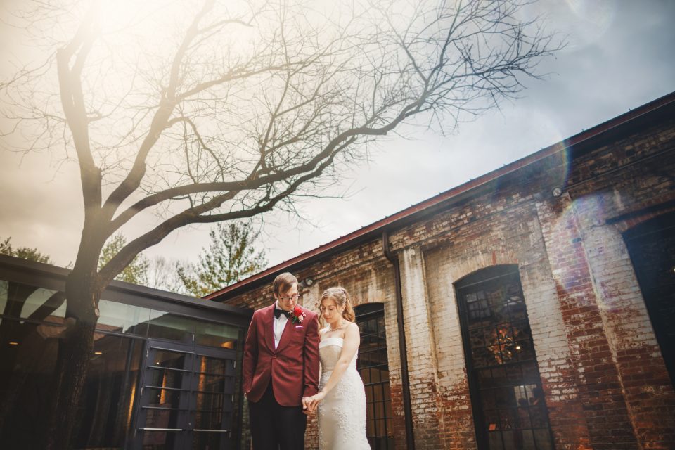A bride and groom stand holding hands outdoors near a bare tree, with an old brick building in the background.