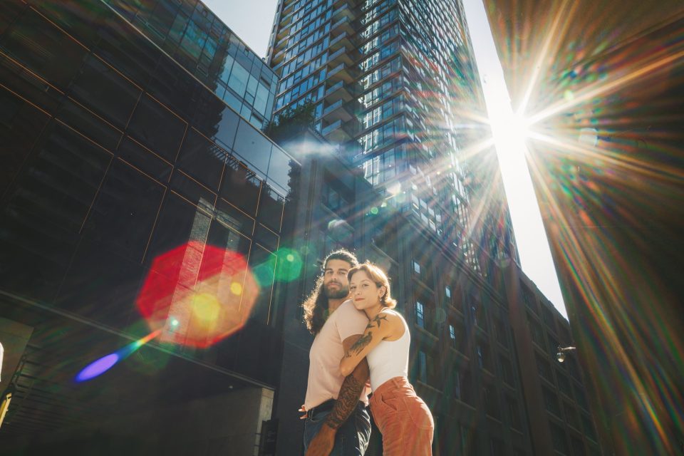 A couple embraces in a city street with tall buildings and a bright sunburst overhead.