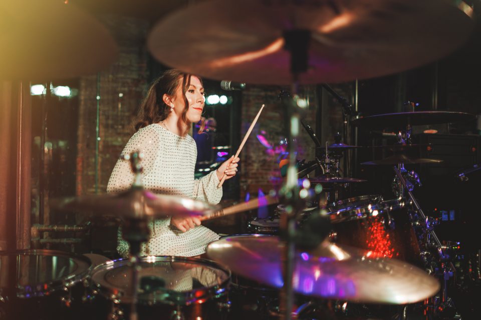 Person playing drums on a dimly lit stage, surrounded by cymbals and drum kits.