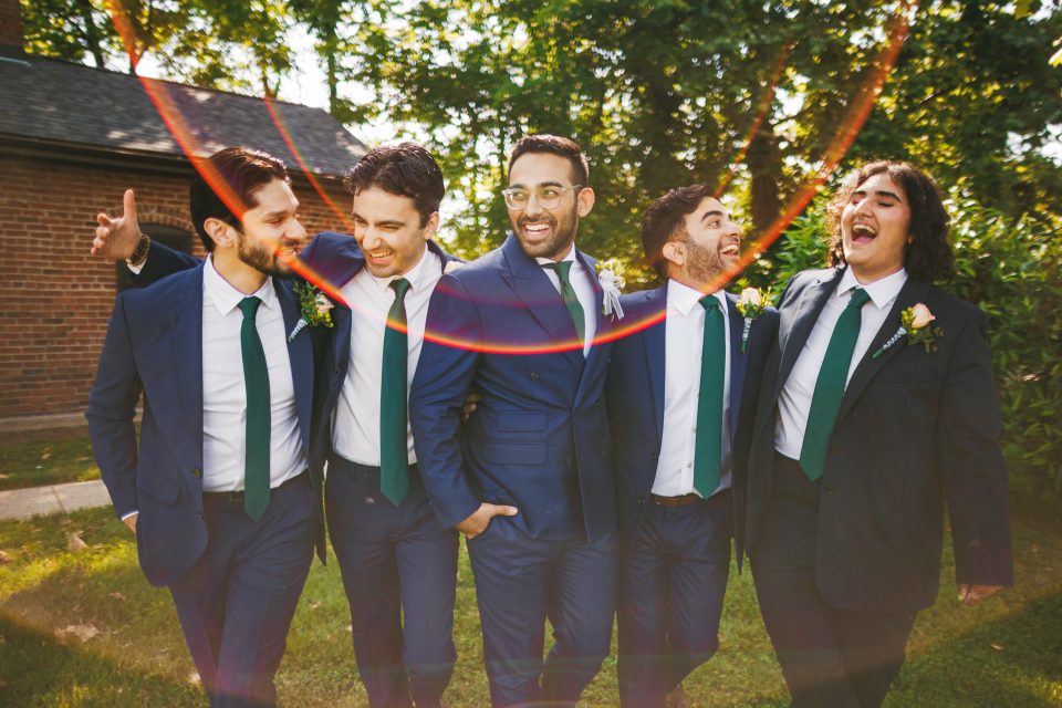 Five men in suits and ties stand together outdoors, smiling and laughing. Sunlight creates a red lens flare across the image.