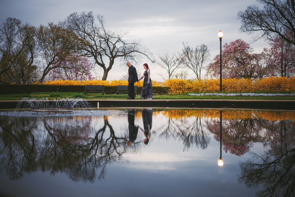 A couple holding hands walks by a pond with their reflection visible, surrounded by blooming trees and yellow flowers under an overcast sky.
