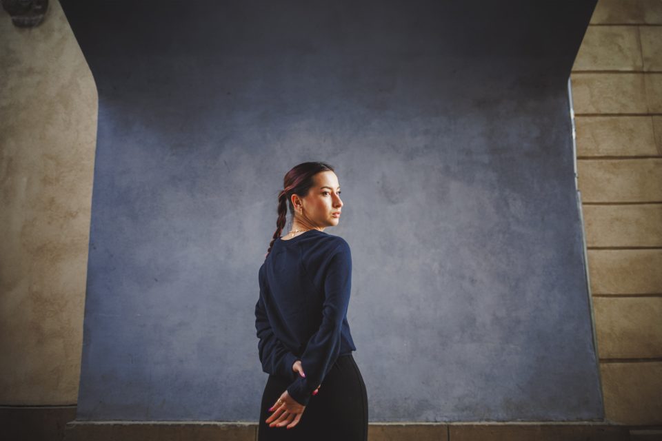 A woman with braided hair stands in front of a textured blue and beige wall, looking over her shoulder with her hands behind her back.
