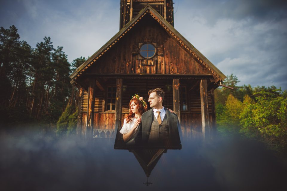 A couple in wedding attire stands in front of a rustic wooden church, with their reflection visible in a body of water below.