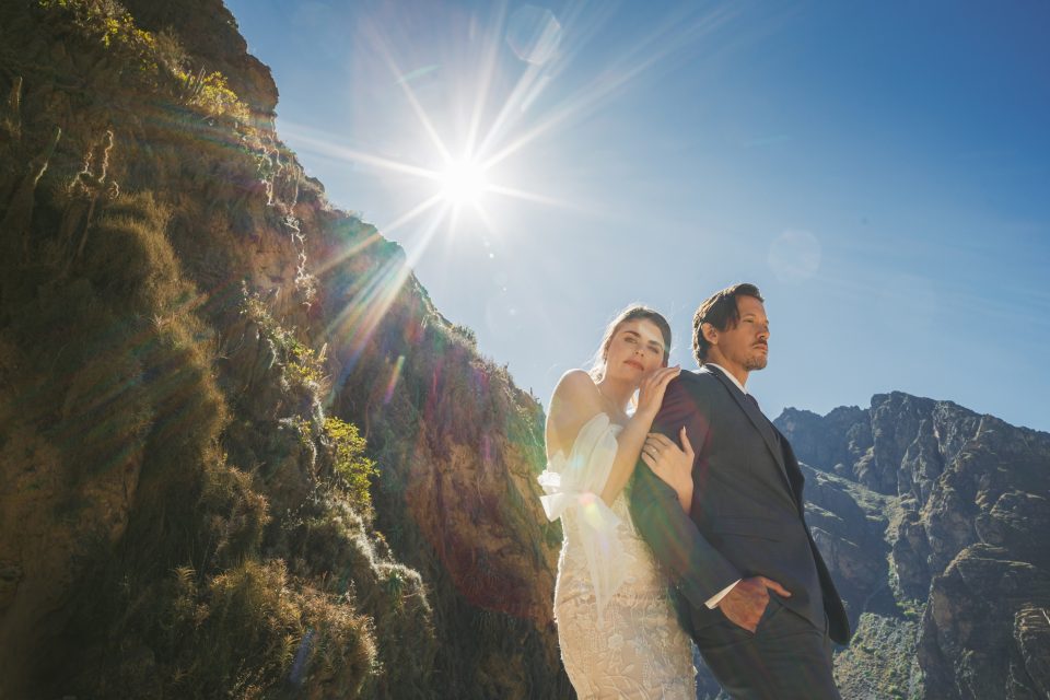 A couple in formal attire stands against a rocky mountain backdrop with the sun shining brightly overhead.