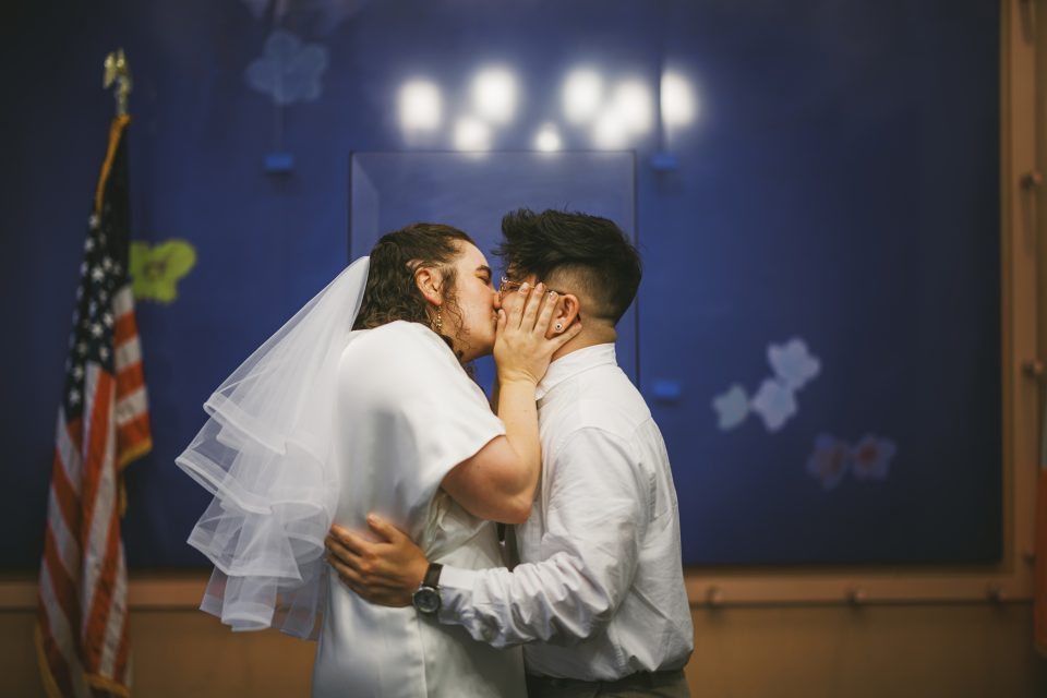 Two people kissing each other while holding hands on their faces, with an American flag and a blue wall in the background. They are wearing white attire.