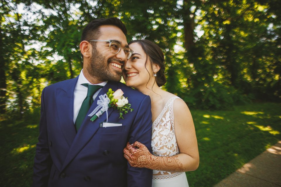 A couple in wedding attire smiles happily outdoors. The groom wears a navy suit with a boutonniere, and the bride in a white dress has henna on her hand. They are surrounded by greenery.