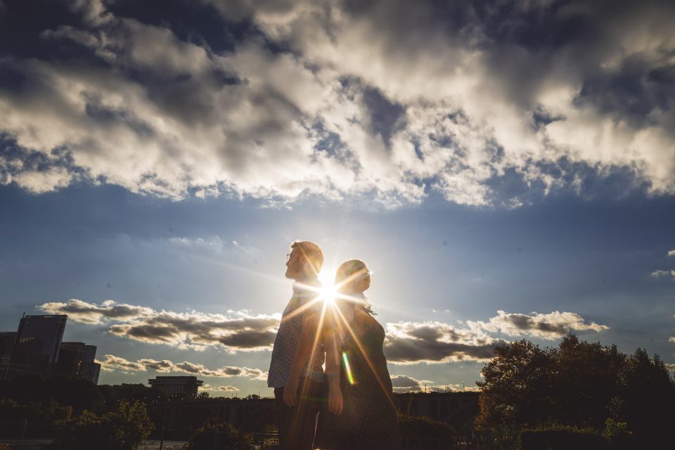 Two people standing back to back with the sun shining brightly between them, surrounded by a partly cloudy sky and distant buildings.