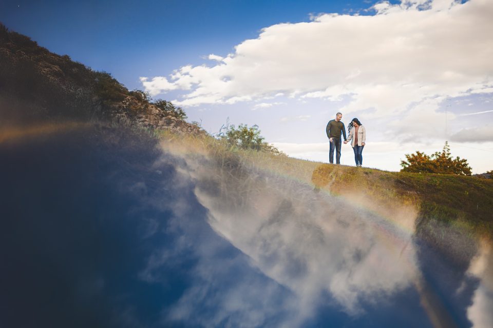 A couple stands on a grassy hill under a partly cloudy sky, with a faint rainbow reflected below them.
