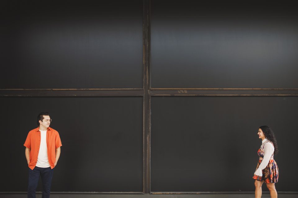 A man in an orange shirt and a woman in a dress stand apart, facing each other, against a large black wall.