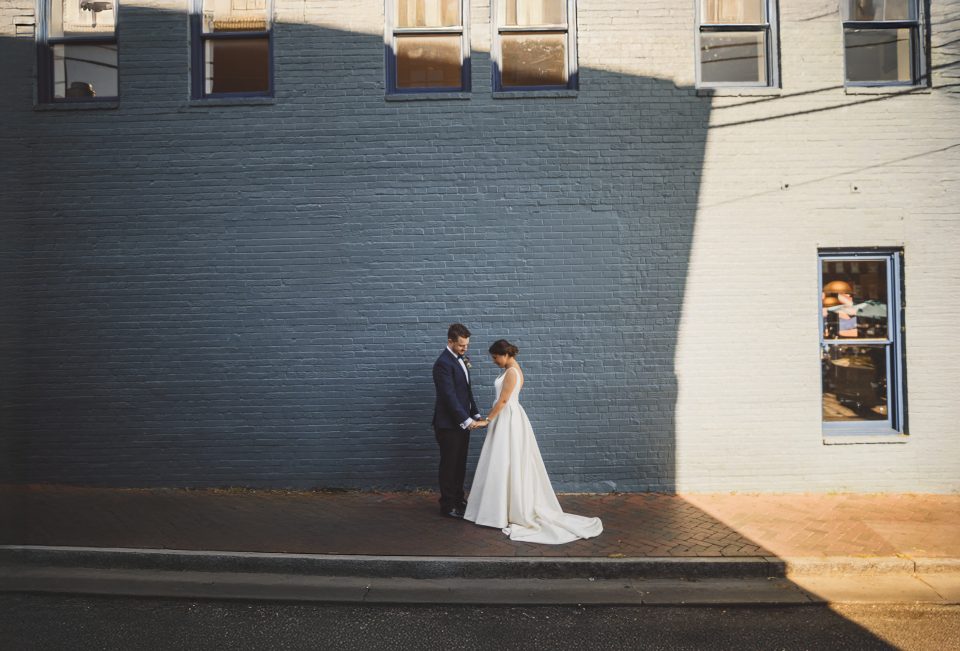 A bride and groom stand together on a sidewalk, holding hands, in front of a large brick building with sunlight casting a shadow.