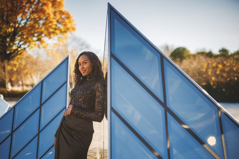 A woman in a black dress leans against a geometric blue structure outdoors on a sunny day, with autumn trees in the background.