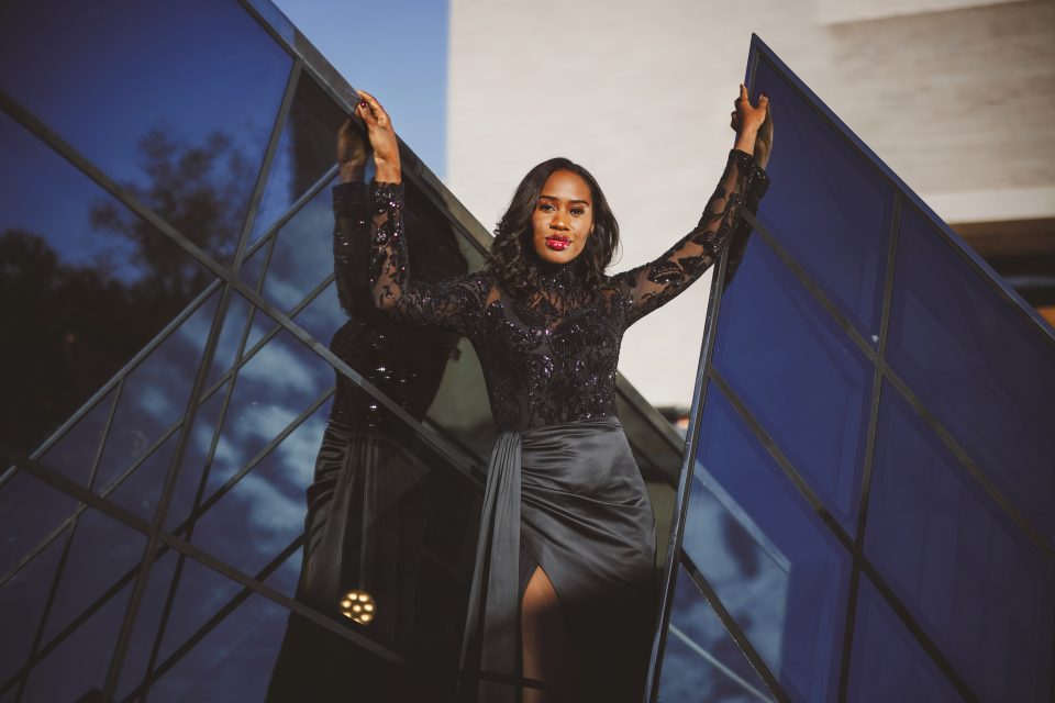 A person in a black dress stands confidently with arms raised, leaning against a modern glass structure, reflecting sunlight.