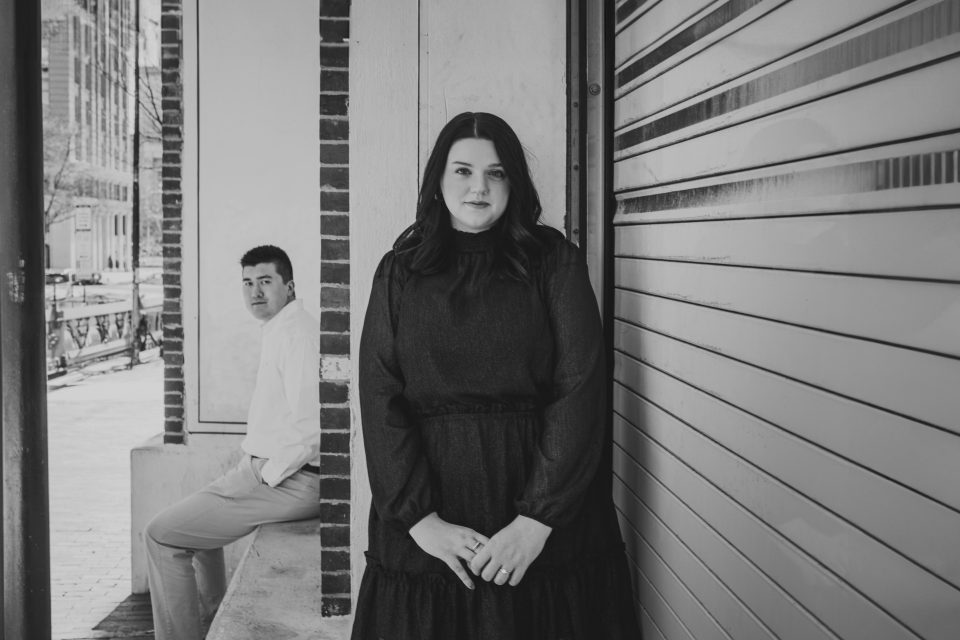 A woman in a dark dress stands near a wall, looking at the camera. A man in a white shirt and light pants sits in the background, looking towards the street.