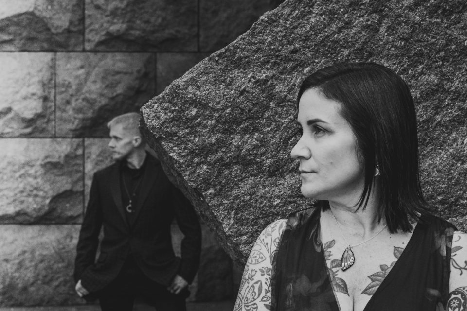 Woman with tattoos and a man in black stand apart in front of a stone wall.
