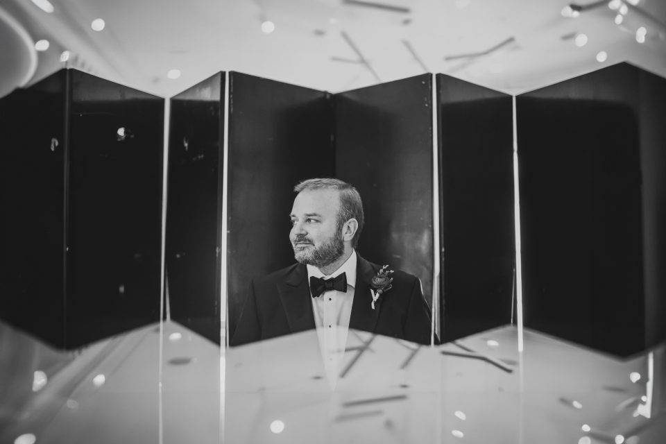 A man in formal attire with a bow tie stands in front of a geometric, reflective backdrop, looking to the side. The image is in black and white with light reflections around him.