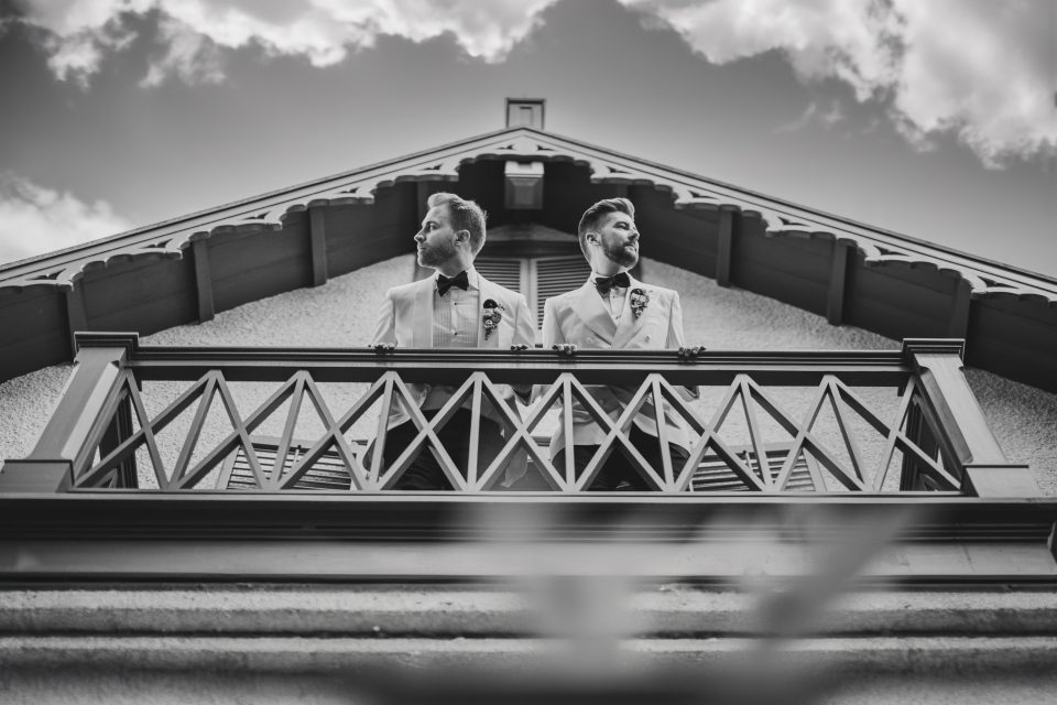 Two people in formal attire stand on a balcony, facing opposite directions, under a cloudy sky. The balcony features intricate woodwork.