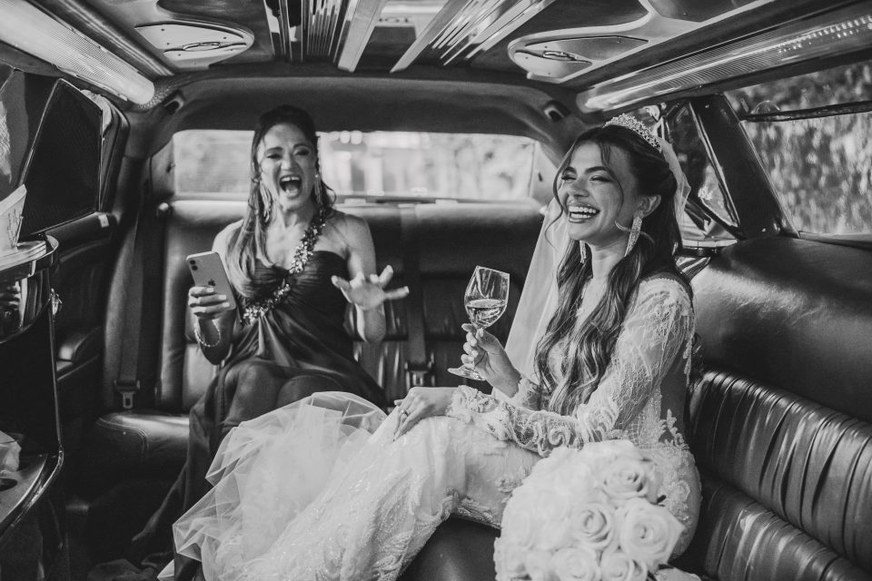 Bride in a wedding dress laughs while holding a wine glass next to a woman in a formal gown, both seated inside a limousine.
