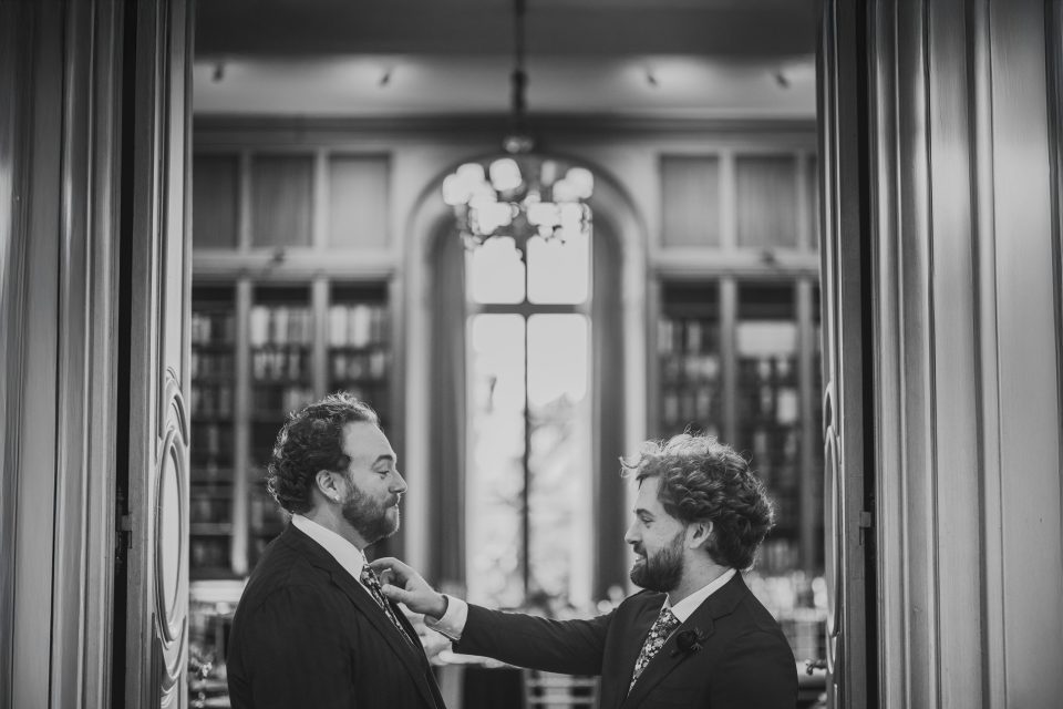 Two men in suits stand facing each other in a formal room with shelves and a large window. One adjusts the other's tie. The scene is captured in black and white.