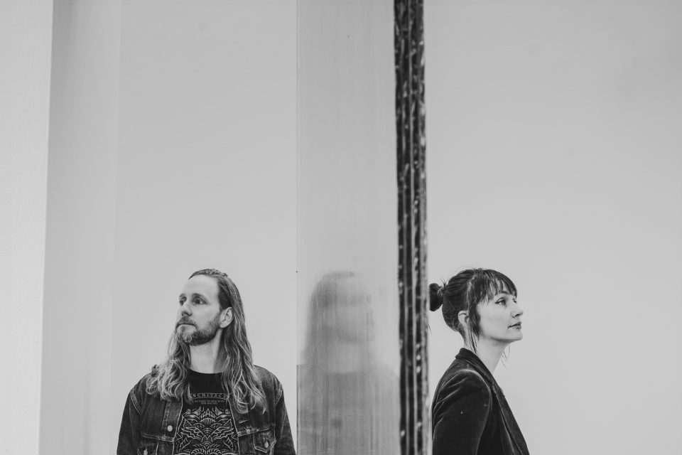 A man and a woman stand on opposite sides of a reflective surface, facing opposite directions, in a monochrome setting.