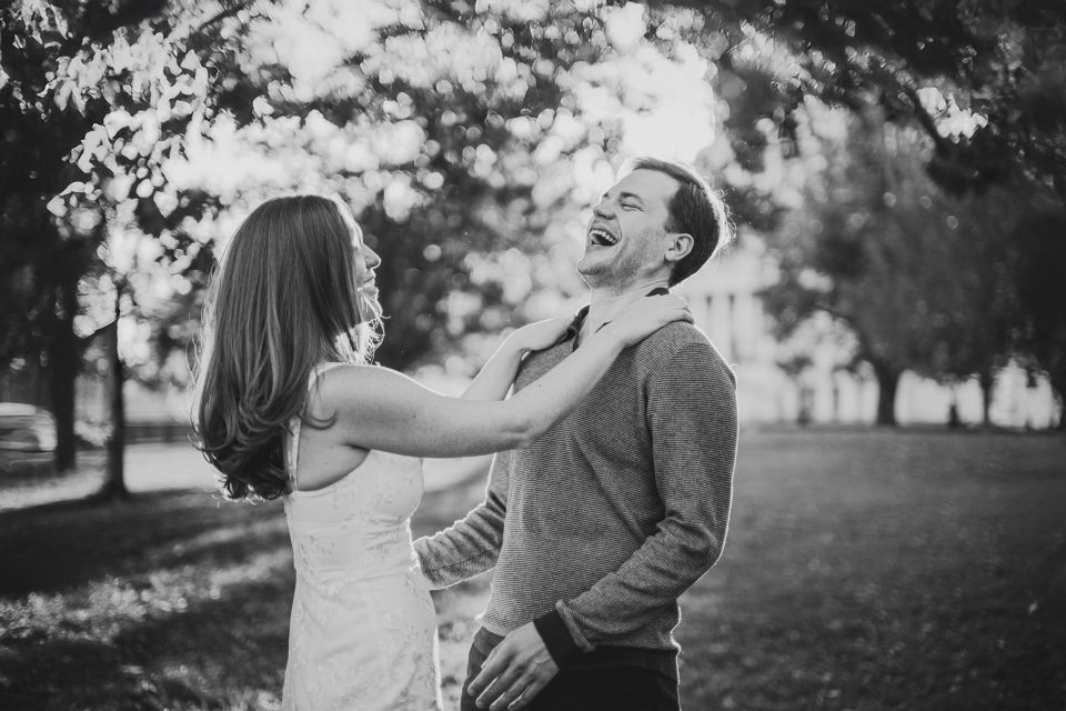 A woman and a man share a lighthearted moment, laughing together in a park. Black and white photo with trees in the background.
