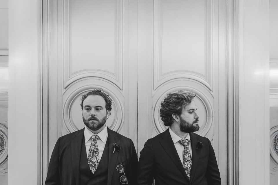 Two men in suits standing back-to-back against ornate double doors. Both wear floral ties, one holds sunglasses. Black and white image.
